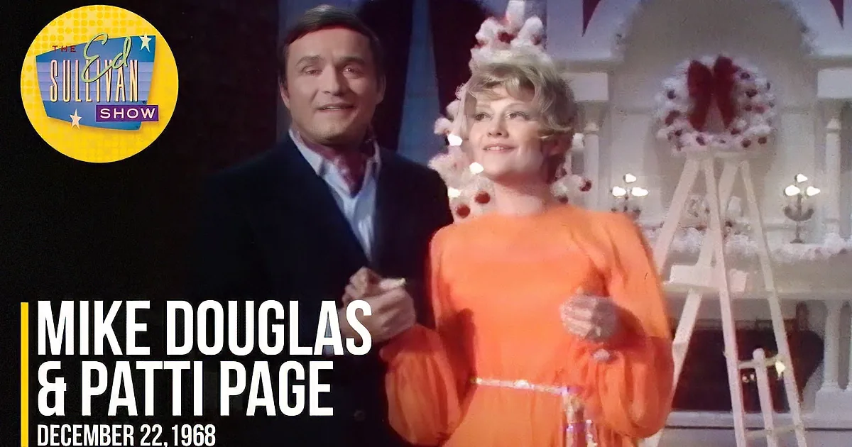 A man in a suit and a woman in an orange dress stand together, smiling, with a decorated Christmas tree in the background.