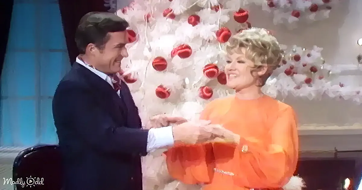 A man in a suit and a woman in an orange dress are holding hands, smiling, with a Christmas tree in the background.