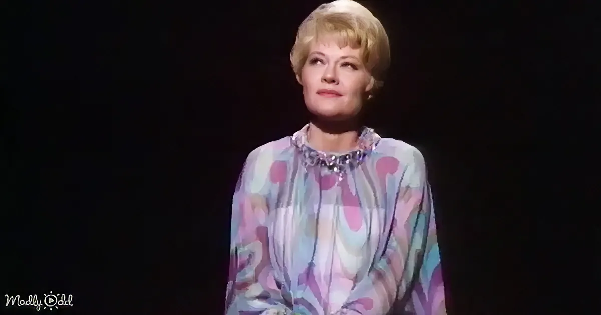 A woman with blonde hair in a colorful, patterned blouse stands against a dark background.