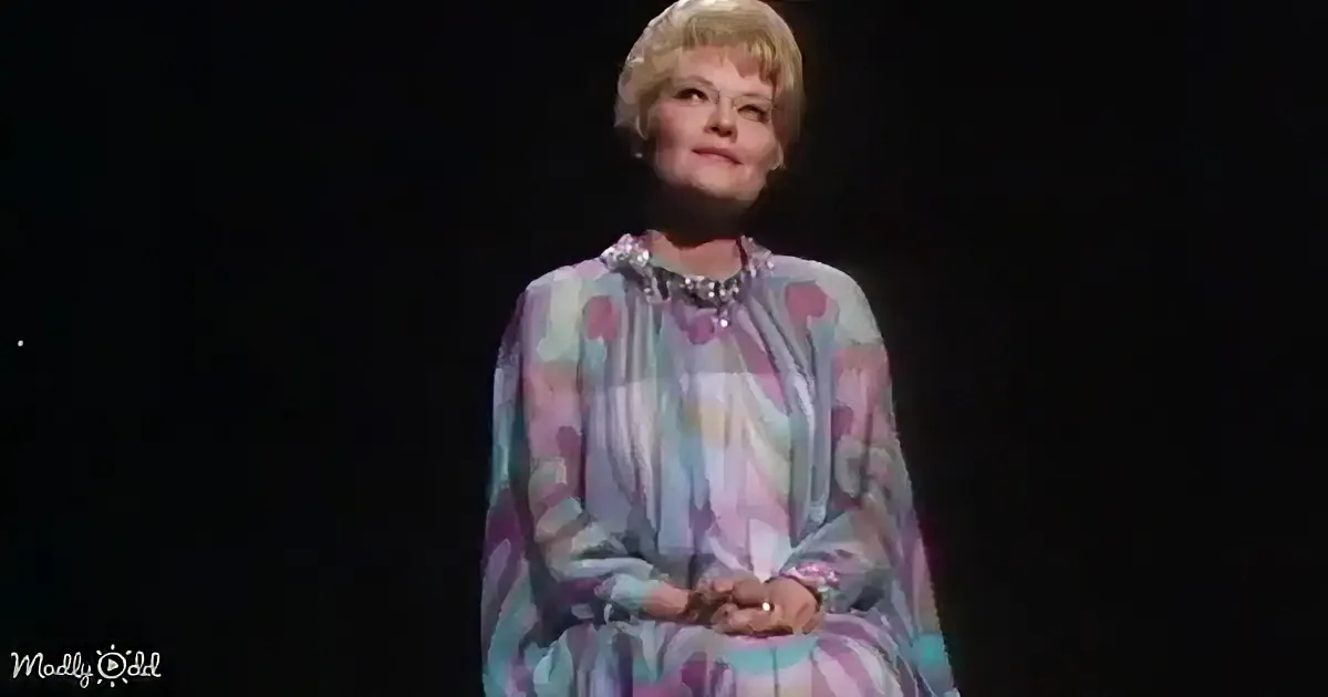 A woman in a colorful polka-dot dress stands on stage, hands clasped, against a dark background.