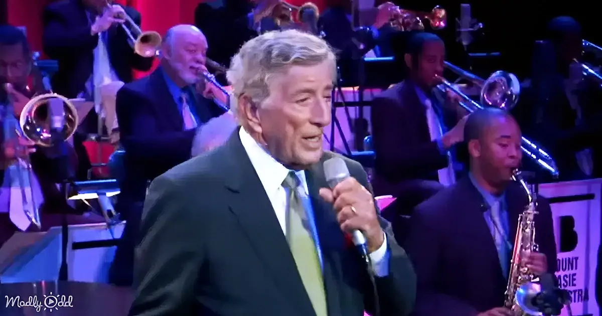 A man in a suit sings into a microphone, surrounded by musicians playing brass and woodwind instruments.