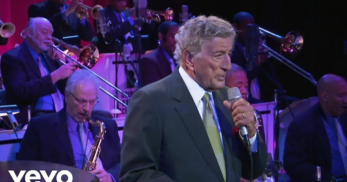 A man in a suit sings into a microphone on stage, with another man playing piano in the background.