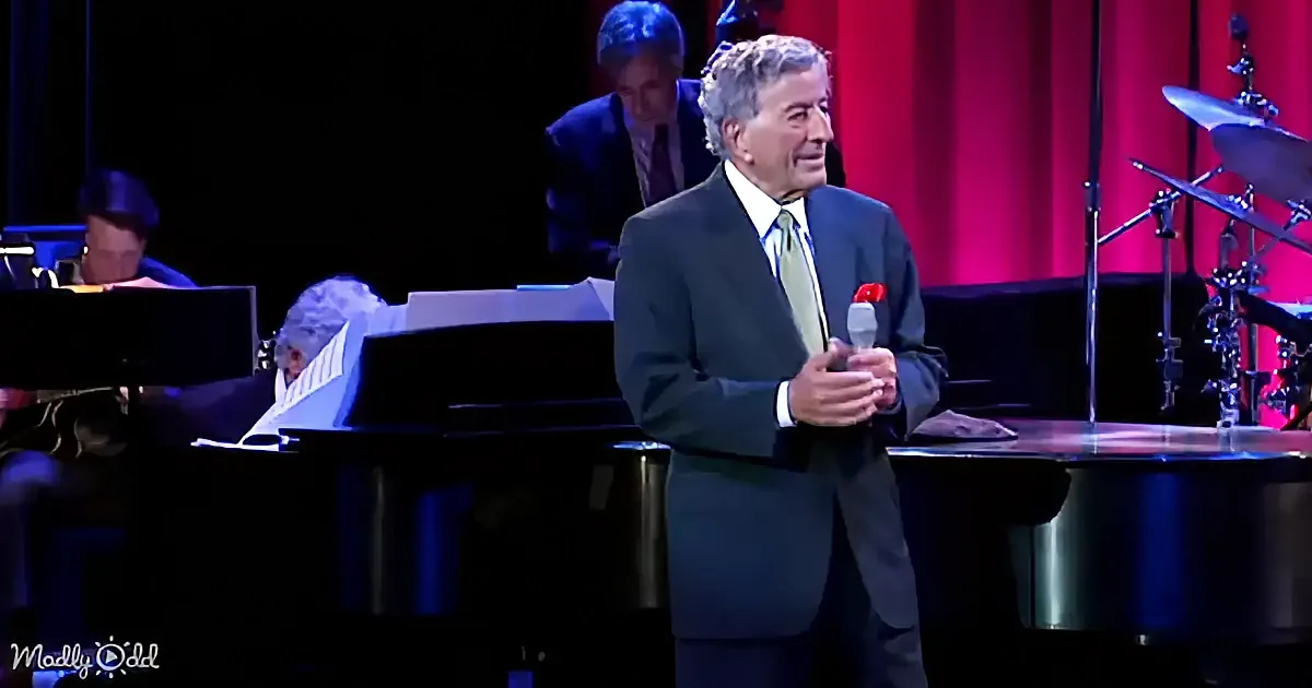 A man in a suit stands on stage holding a microphone, with a piano and drum set in the background.
