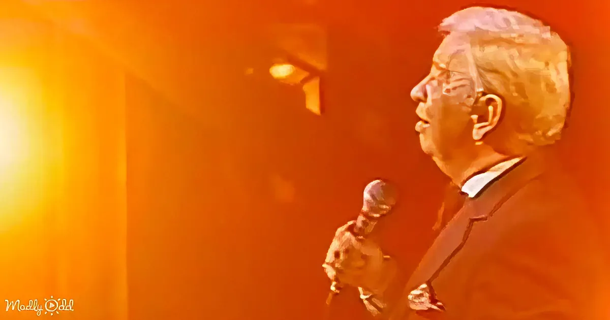 A man in a suit holds a microphone, speaking against a warm, orange-lit background.