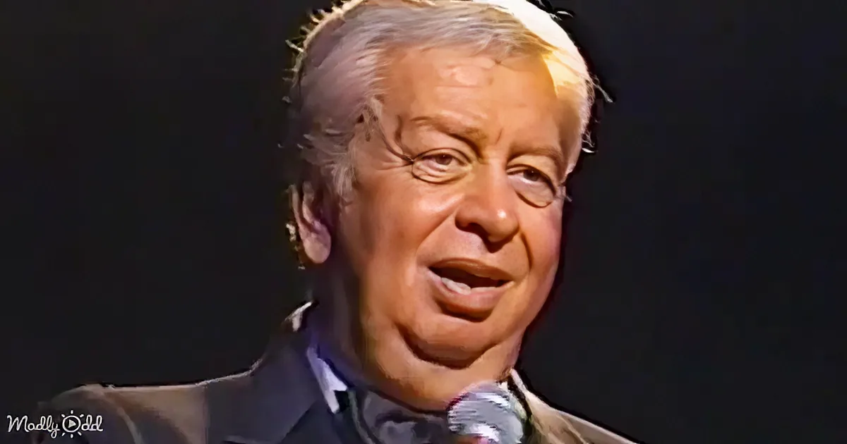 A man in a suit speaking into a microphone, with a dark background and a watermark in the corner.