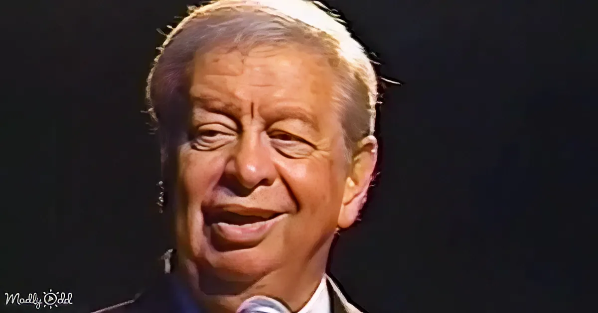 A smiling man with short, light-colored hair and a microphone, wearing a dark suit and white shirt, against a dark...