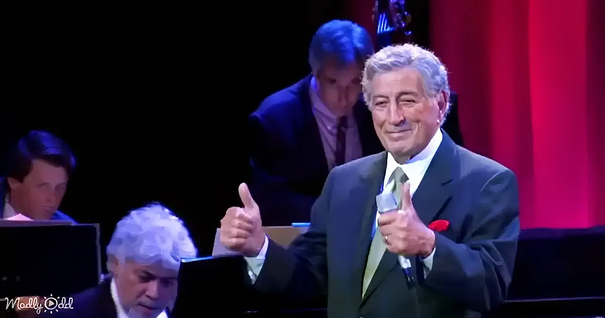 A man in a suit gives two thumbs up on stage, smiling, with musicians in the background.