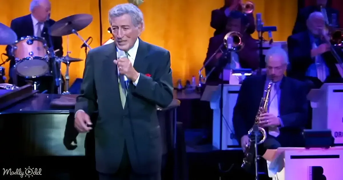 A man in a suit sings into a microphone on stage with a band playing behind him.