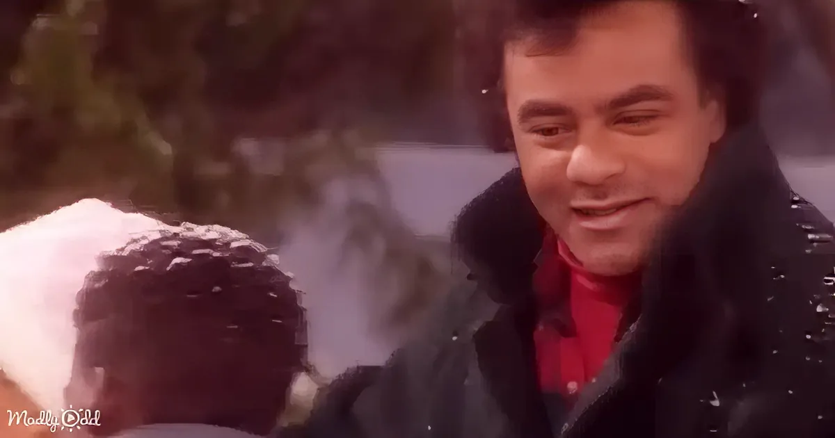 A man in a black coat and red shirt smiles, snow on his hair, with another person in the background.