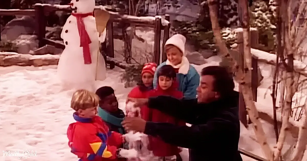 A man in a black jacket holds a snowball, surrounded by children in winter gear, with a snowman in the background.