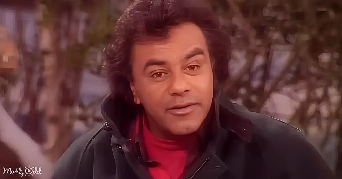 A man with dark hair, wearing a black jacket over a red shirt, stands outdoors with trees in the background.