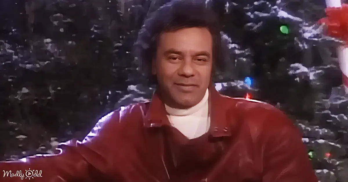 A man in a red jacket smiles in front of a Christmas tree with lights.