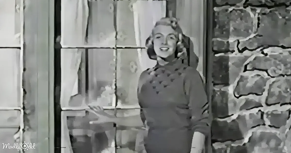 A smiling woman in a patterned dress stands near a stone wall and window, gesturing with her hand.