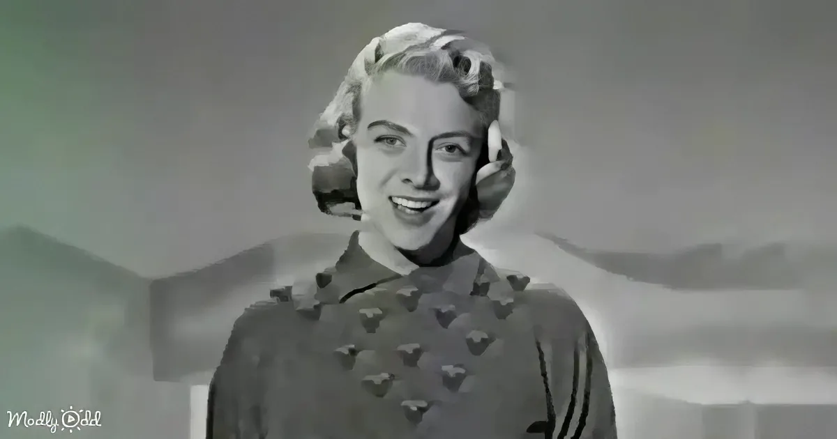 A smiling woman with short, styled hair, wearing a patterned dress, stands against a muted background.