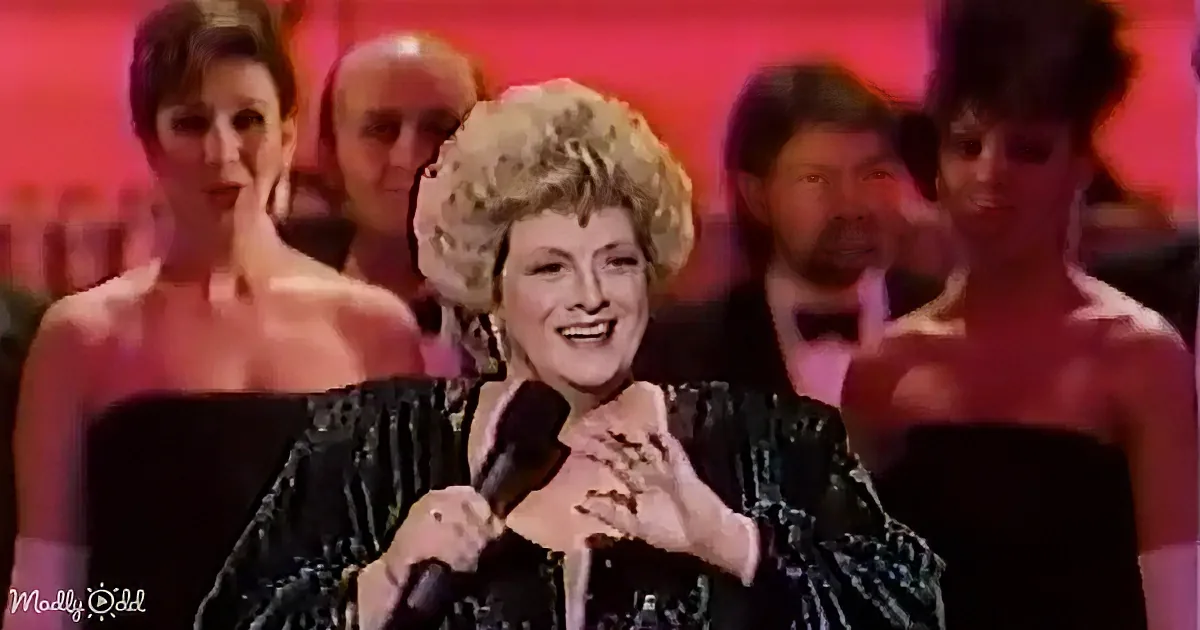 A woman in a black sequined dress holds a microphone, smiling, with others in the background.