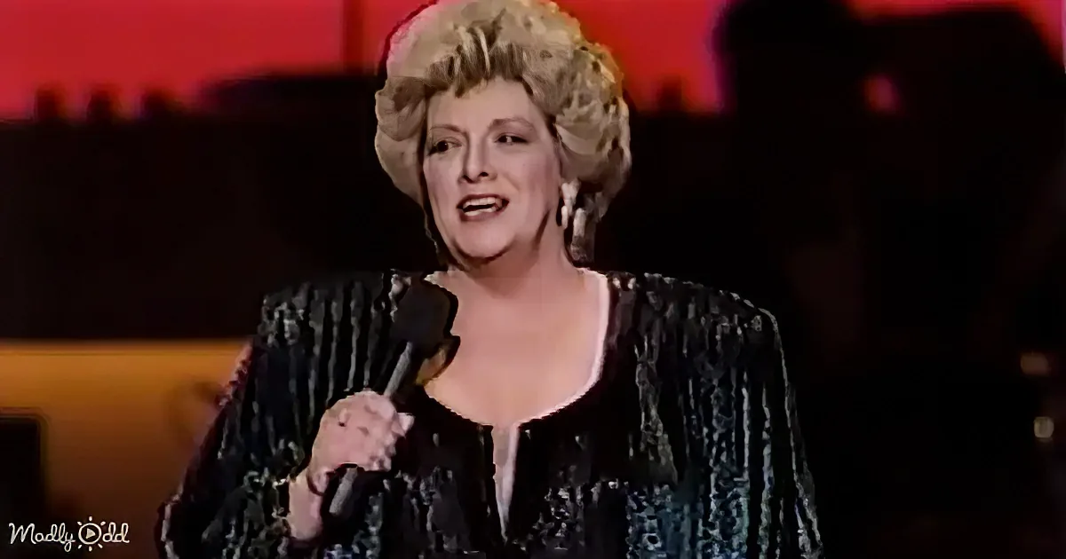 A woman with blonde hair, wearing a black sequined outfit, sings into a microphone on stage.