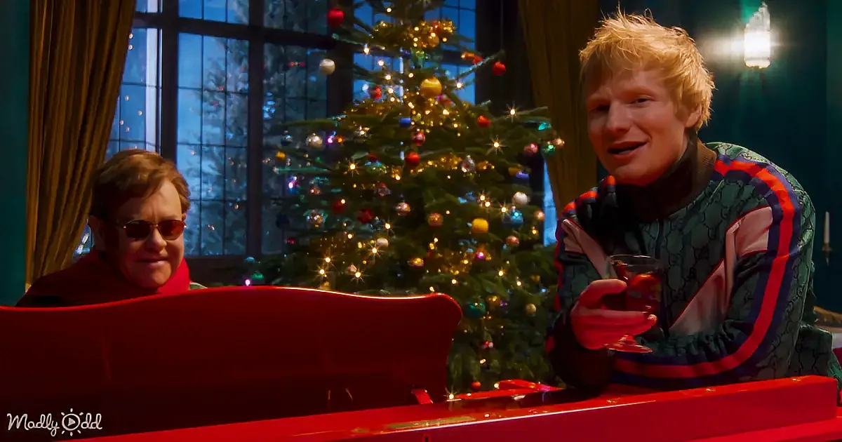 Ed Sheeran holds a drink, smiling, with Elton John playing piano in front of a decorated Christmas tree.