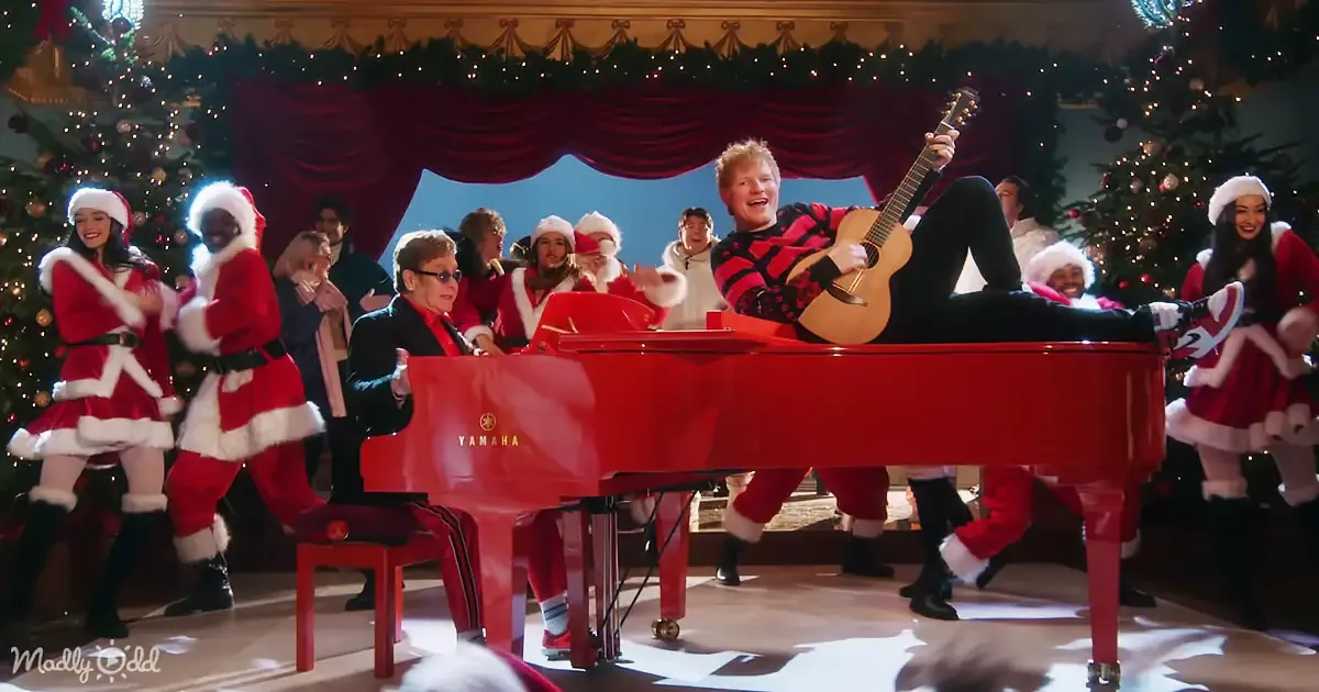 Ed Sheeran plays guitar on red piano, surrounded by dancers in Santa costumes, festive Christmas setting.