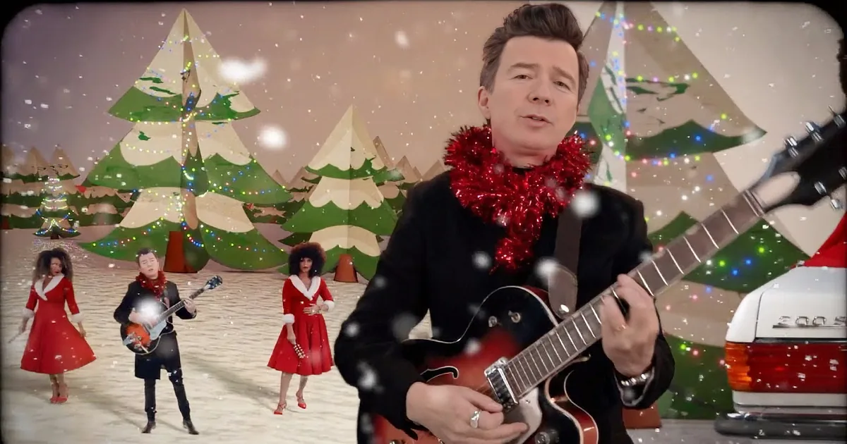 A man plays guitar on a white car surrounded by dancers and a snowman in a festive Christmas setting.