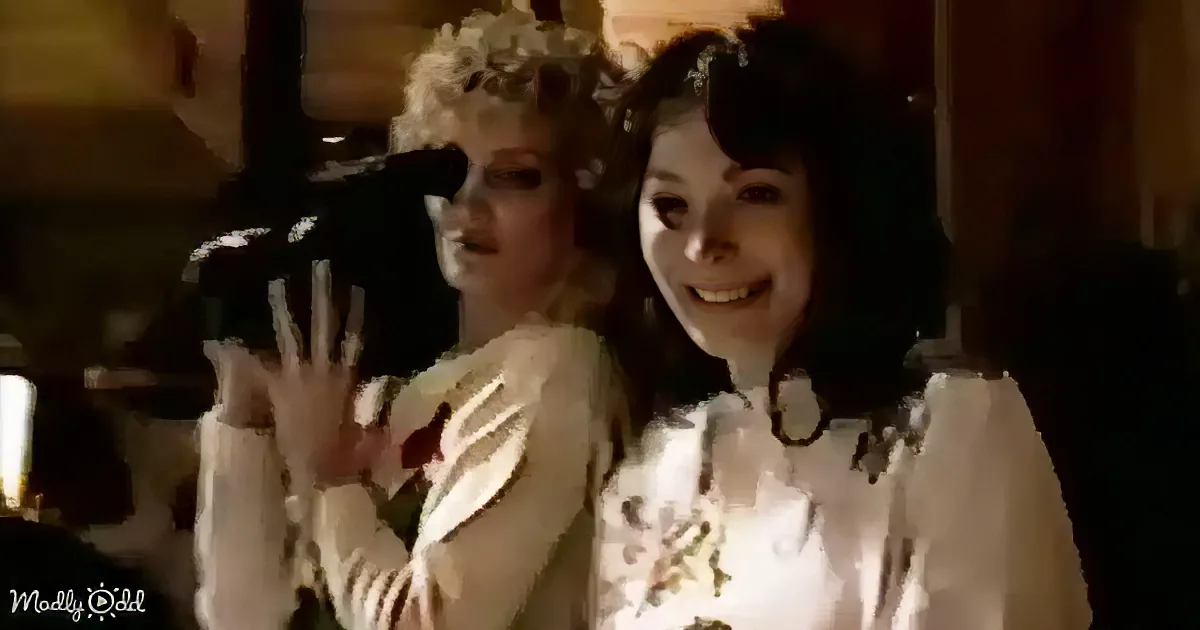 Two women in white outfits, one smiling, the other gesturing with hands, in a warmly lit room.