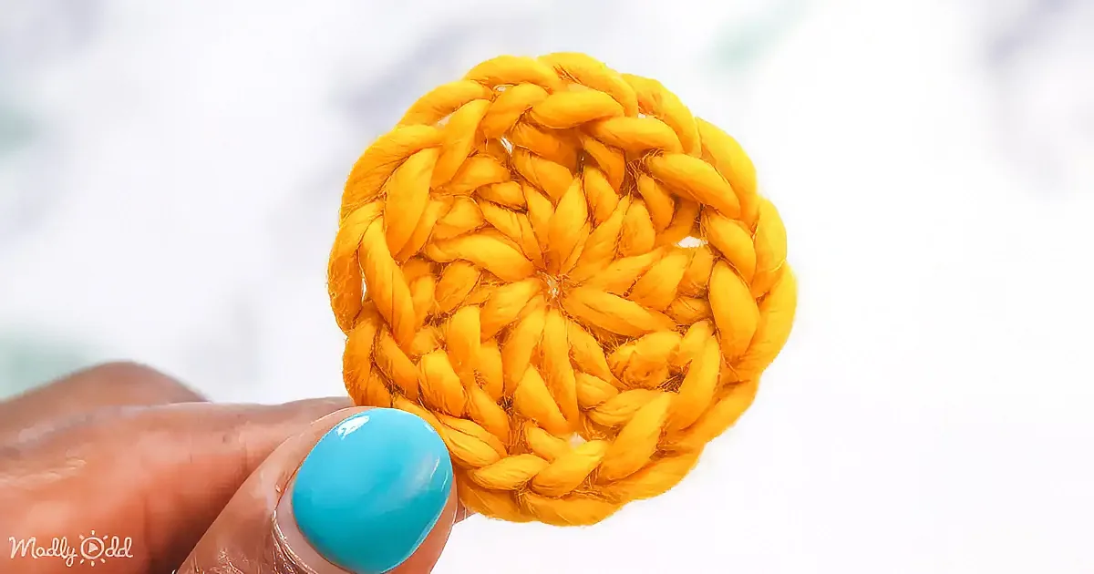 A hand holds a circular, yellow crocheted item with a textured pattern against a white background.