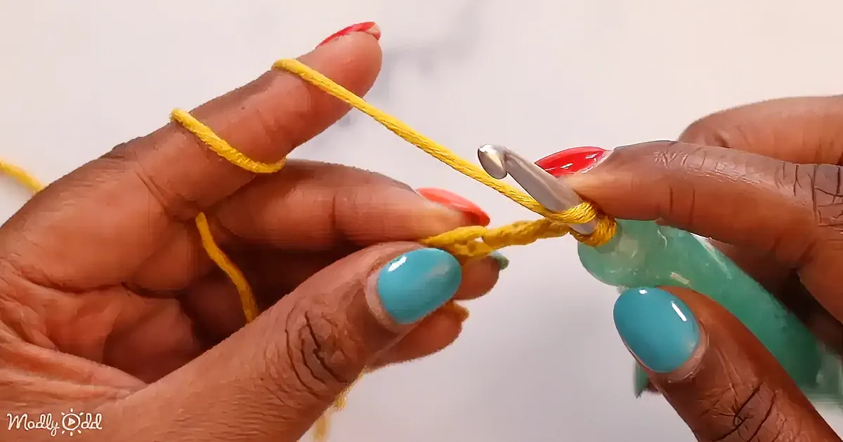 Hands with colorful nails hold a yellow yarn and a crochet hook, demonstrating a crochet technique.