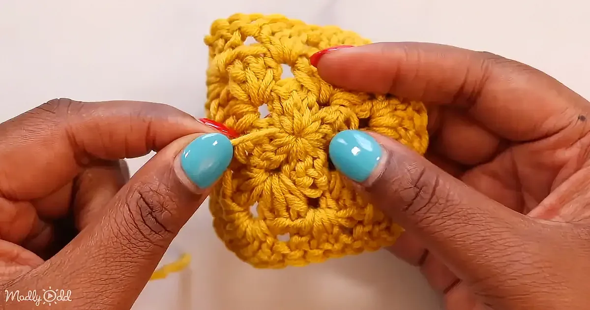 A person with turquoise nails holds a yellow crocheted square with a red crochet hook.