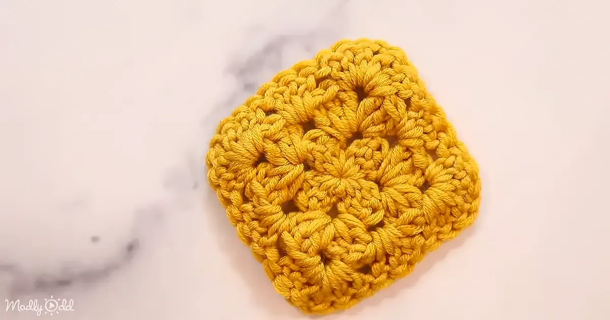 A yellow crocheted hexagon with intricate patterns lies on a white marble surface.