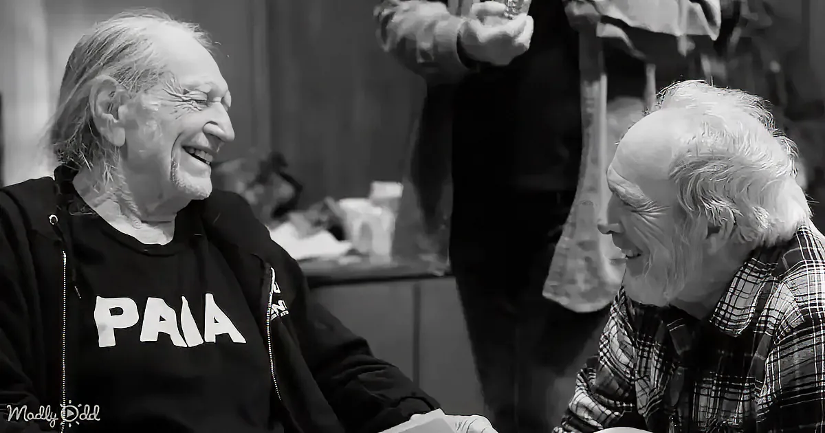 Two older men are laughing and smiling, one wearing a "PAIA" hoodie, in a casual indoor setting.