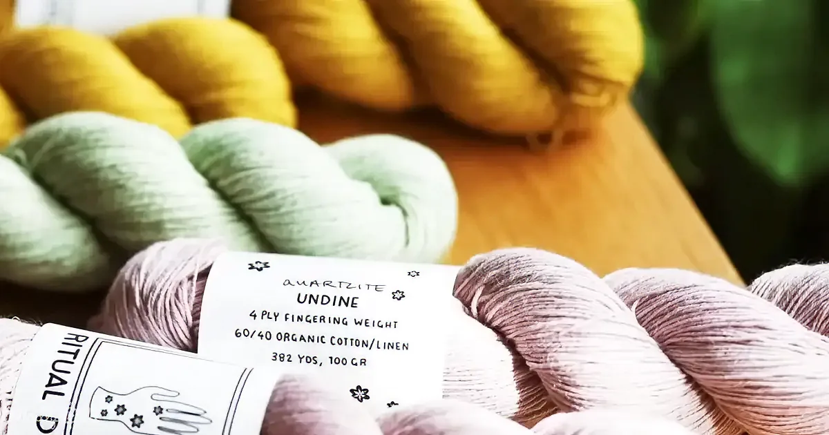 A close-up of colorful yarn skeins, with a focus on a purple skein labeled "Quartzite Undine," showcasing organic...
