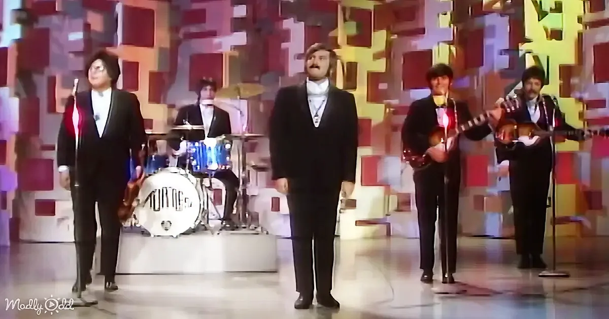 A man in a black suit stands center stage with a band, including a drummer and musicians with instruments.