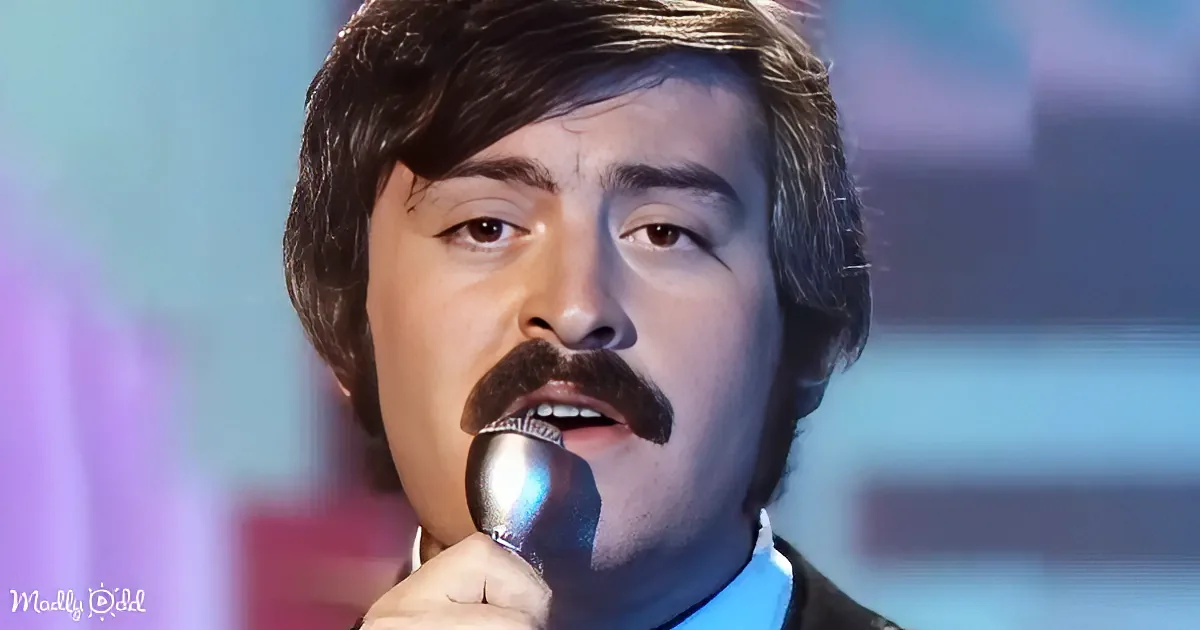A man with a mustache speaks into a microphone, illuminated by green light, against a blurred red background.