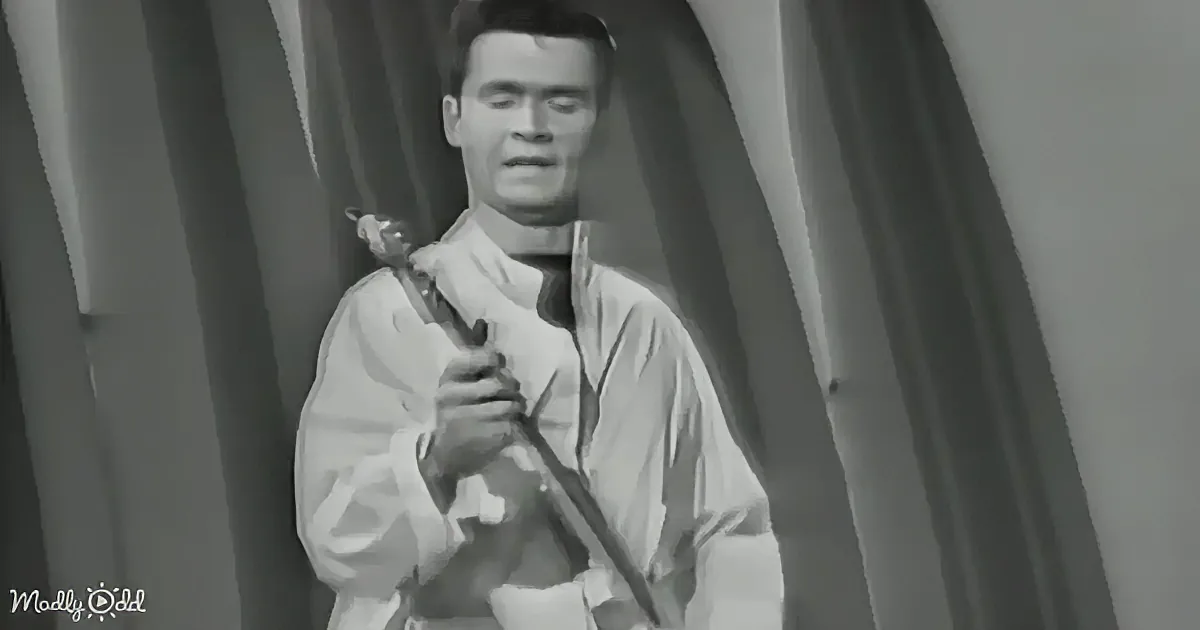 A man in white attire holds a sword, standing in front of draped fabric.