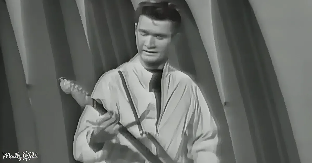 A man in a light-colored jacket holds a guitar, looking down, with a curtain backdrop.