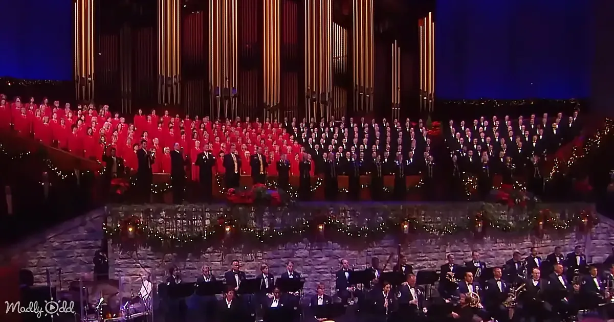 A large choir in red and black attire stands on stage with an orchestra below, both adorned with festive decorations.