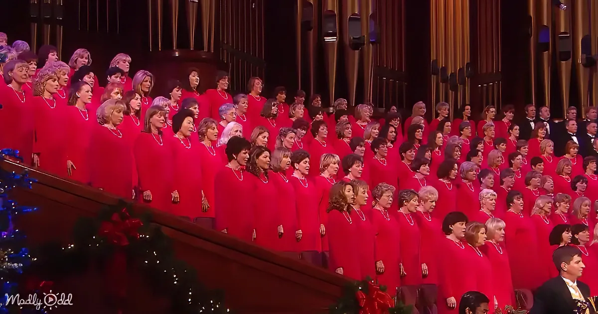 A large choir in red robes sings in a grand hall with organ pipes.