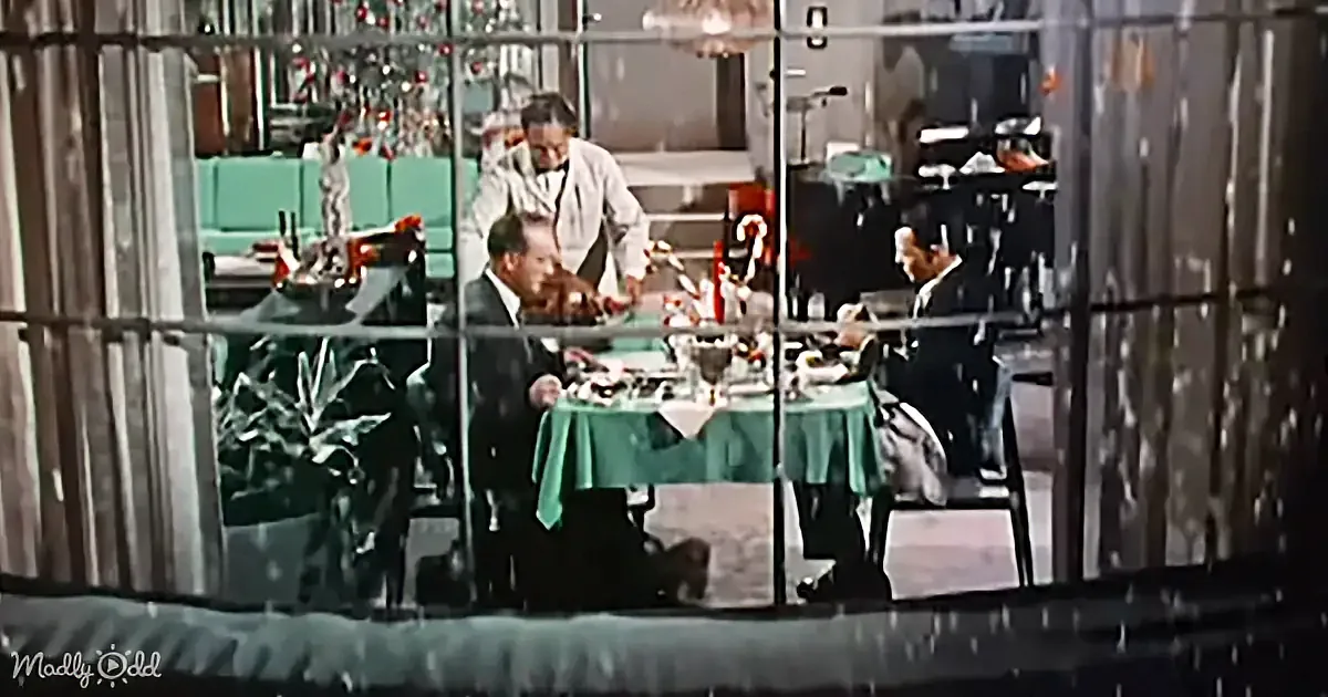 A man in a suit sits at a table with a green tablecloth, surrounded by other people in a festive setting.