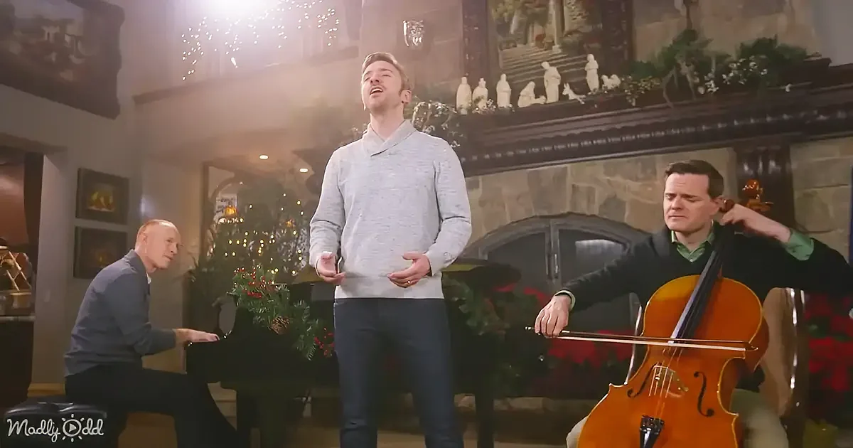A man in a grey sweater sings passionately while another plays the cello and a third plays the piano in a festive setting.
