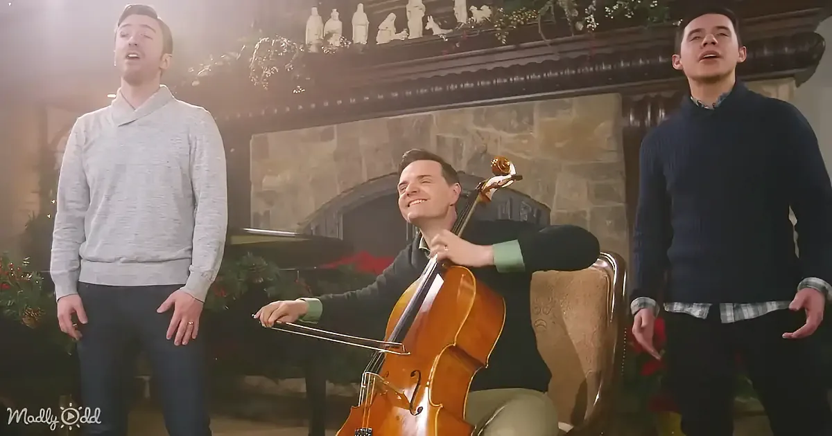 A man plays cello while two others sing in front of a decorated fireplace.