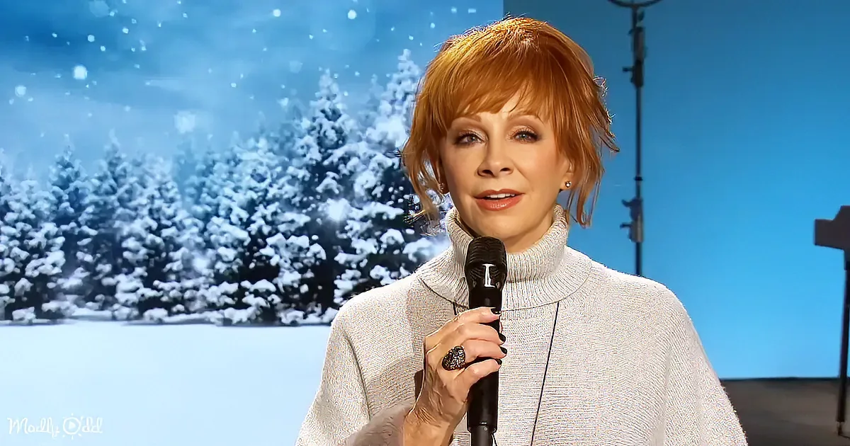 A woman with red hair holds a microphone, wearing a gray sweater, against a snowy backdrop.