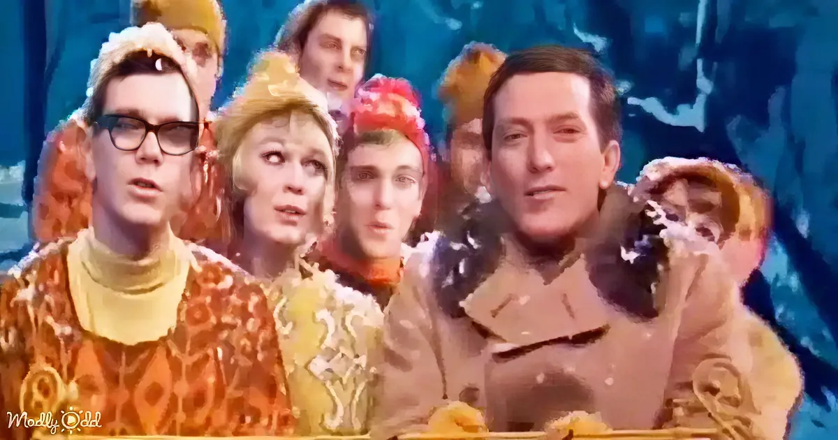 A man in a brown coat sits with a group of people in colorful, patterned outfits against a blue background.