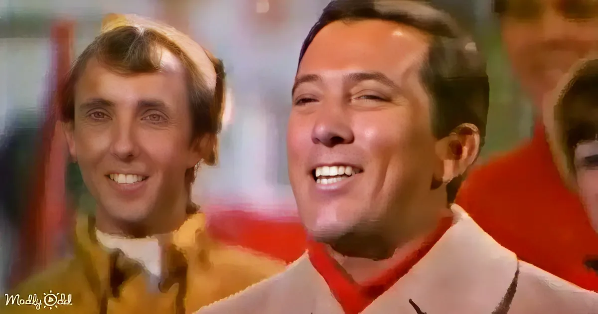 A man in a beige suit and red shirt smiles, with another man in a yellow jacket laughing in the background.