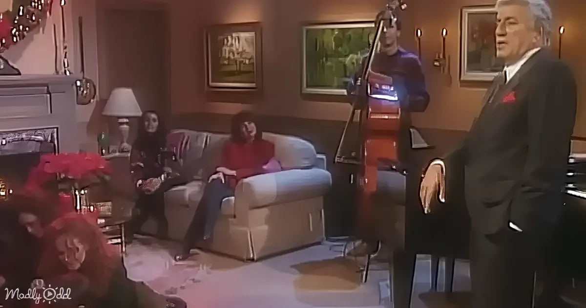 A man in a suit stands in a living room with a cello player and two women on a couch, decorated for Christmas.