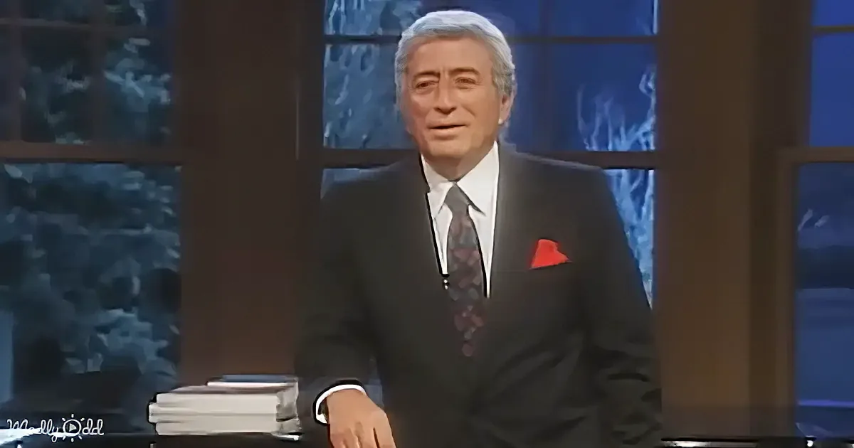 A man in a suit and tie stands in front of a windowed backdrop, appearing to speak or present.