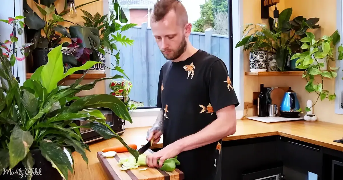 A man with a mohawk and tattoos slices vegetables on a cutting board in a kitchen filled with plants.