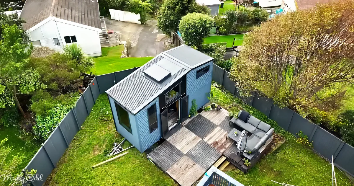 A modern blue shed with a solar panel sits on a wooden deck in a fenced backyard with greenery and a neighboring white house.