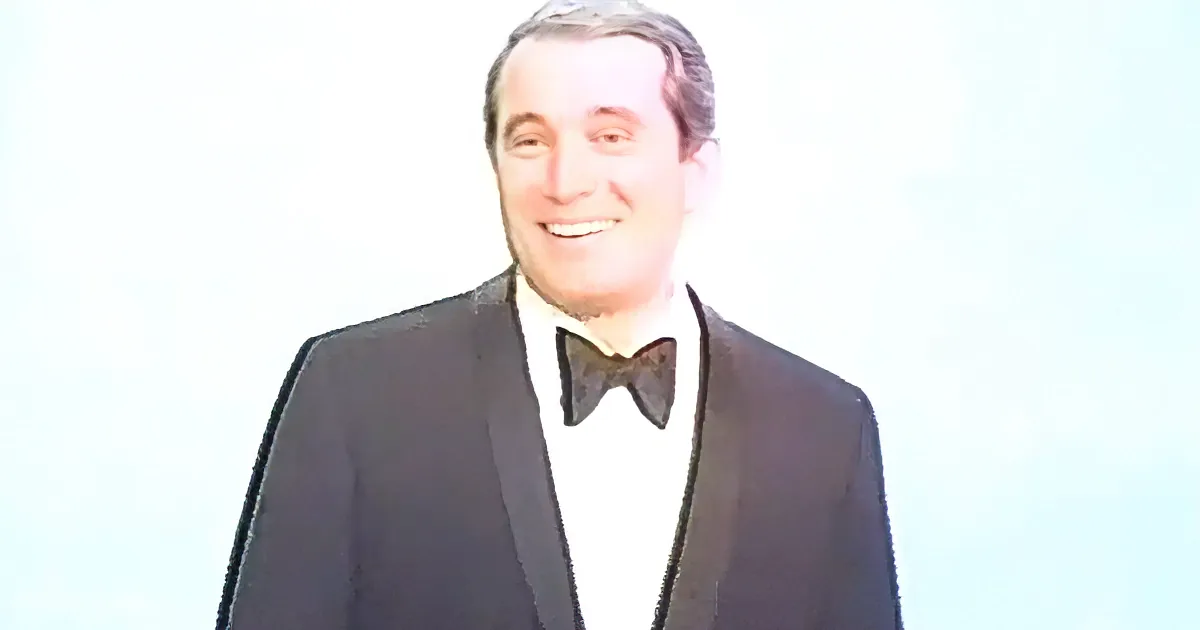 A man in a tuxedo with a bow tie smiles against a light background.
