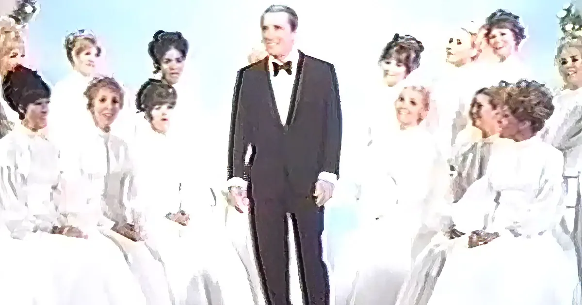 A man in a tuxedo stands in front of a group of women in white dresses.