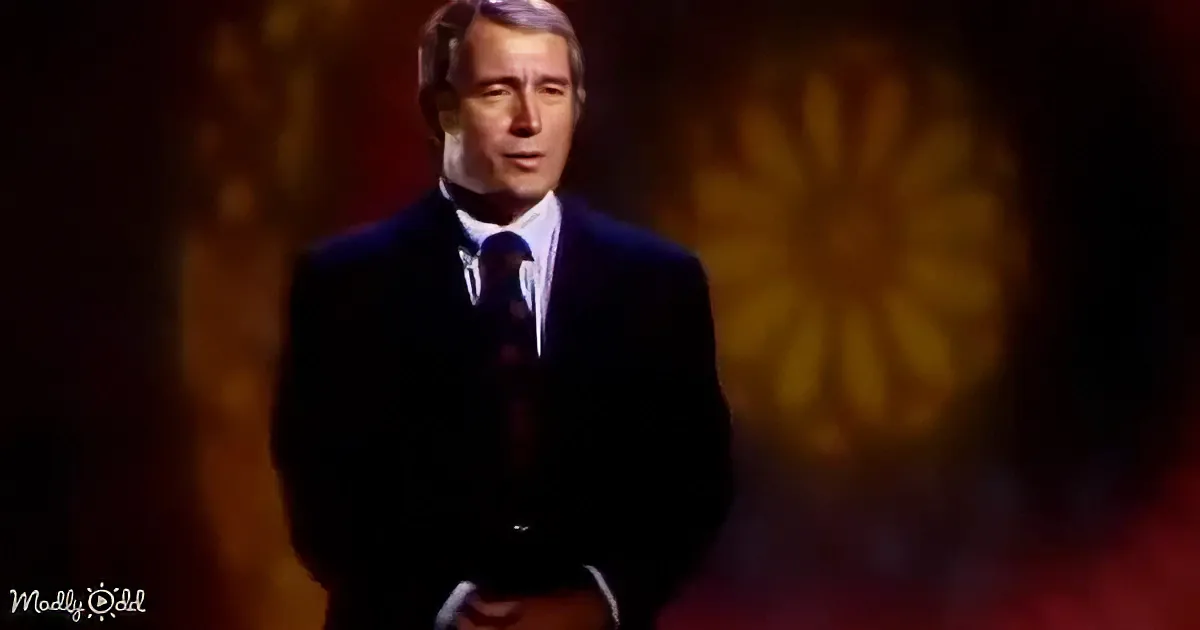 A man in a dark suit and tie stands against a blurred, warm-toned background.