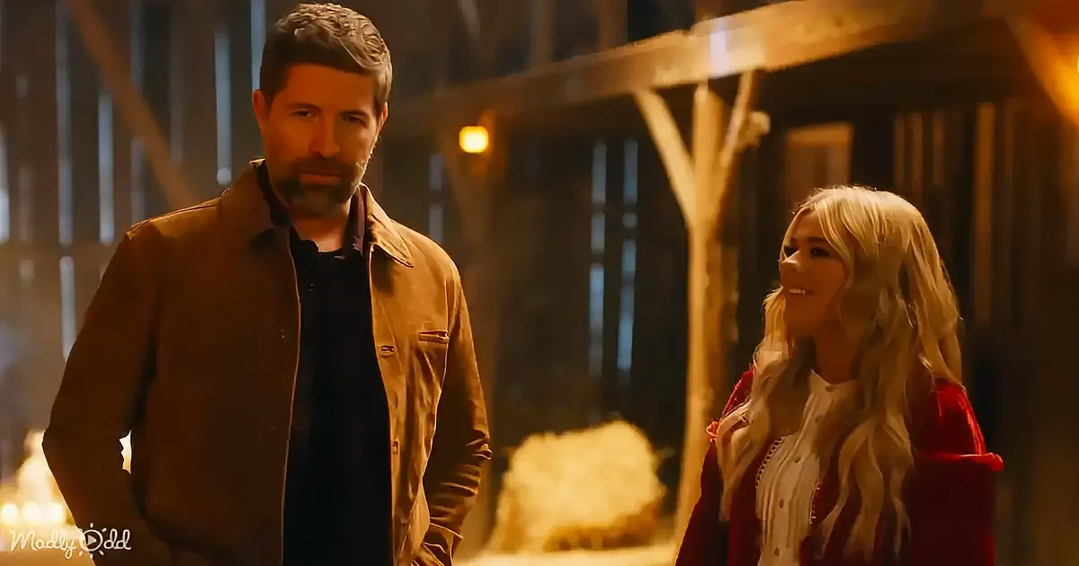 A man in a brown jacket stands next to a woman with long blonde hair in a barn-like setting.